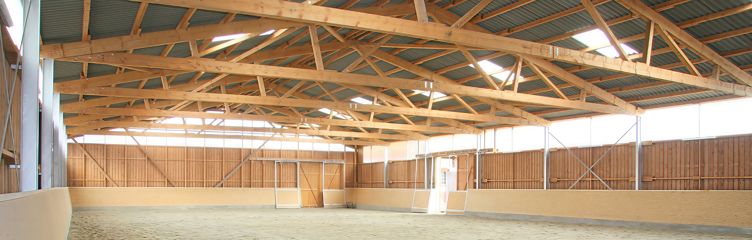 Innenansicht eines Außenklimastalls mit Holztragwerk, großzügigem Lichteinfall und offener Stallstruktur.
